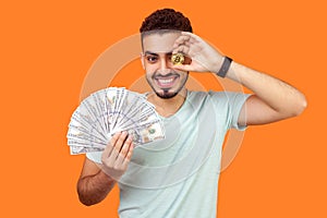 Portrait of joyous rich brunette man holding dollars and covering one eye with golden bitcoin. isolated on orange background