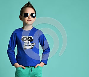 Portrait of kid boy in cool blue aviator sunglasses, blue shirt with print standing with his hands in pockets on gray