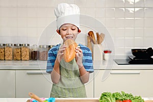 Portrait of kid eating large loaves of bread, happy Asian boy with apron and chef hat holding and eating freshly baked homemade