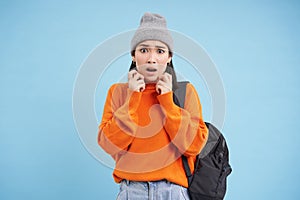 Portrait of korean woman looks scared, jumps terrified, stands with backpack in street clothing, blue background