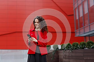 Portrait of a laugh curly young woman, wear in red jacket, read or write messages, over building background.