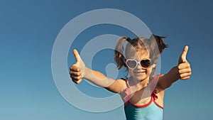 Portrait of laughing girl of 6 years in blue swimsuit and heart-shaped sunglasses gesturing super, fine against blue sky
