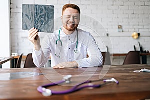 Portrait of laughing male doctor in white uniform examining brain computerized tomography scan sitting at desk in