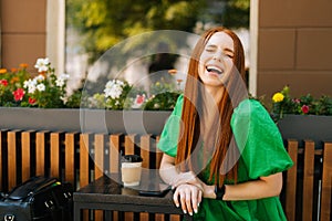 Portrait of laughing redhead young woman sitting at table with coffee cup and mobile phone in outdoor cafe terrace in