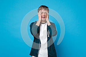 Portrait of laughing young man in casual clothes keeping eyes closed, putting hands on face isolated on blue wall