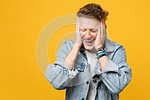 Portrait of laughing young man in denim casual clothes keeping eyes closed, putting hands on head isolated on yellow