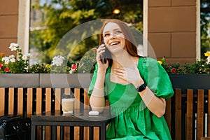 Portrait of laughing young woman talking by mobile phone, sitting at table with coffee cup in outdoor cafe terrace in