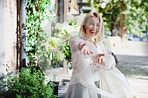 Portrait of Laughing young woman in white trench coat having fun on street. Dancing happy blonde girl pull hands forward.