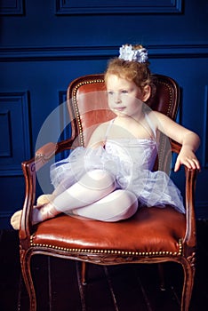 Portrait little 4 years old blonde curly ballerina sitting in dark room on brown leather chair in ballet dress white tutu and