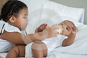 Portrait of little adorable African older sister feeding newborn baby milk bottle with love and care while infant crying lying