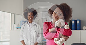 Portrait of little african-american girl holding teddy bear with young female doctor standing on background