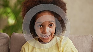 Portrait little African American girl looking at the camera and laughing cheerfully. Teenage girl sits on gray sofa in