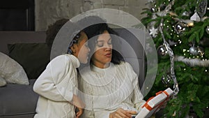 Portrait of a little African American girl surprises her mother with a gift. Mother and daughter sitting near sofa and