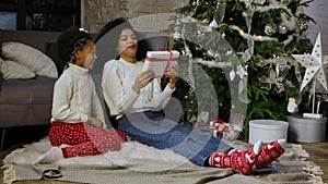 Portrait of a little African American girl surprises her mother with a gift. Mother and daughter sitting near sofa and