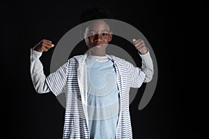 Portrait of little African boy raising his arms and stretching muscles to show his strength with serious face on black background