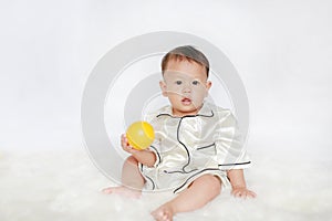 Portrait of little Asian baby boy in pajamas holding yellow plastic toy ball sitting on white fur against white background