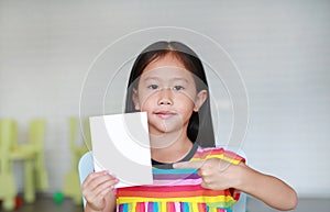 Portrait little Asian child girl holding blank white paper card in her hand with pointing. Kid showing empty paper note copy space