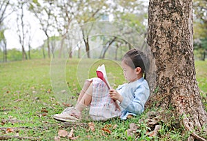 Portrait little Asian child girl reading book in park outdoor sitting lean against tree trunk in the garden outdoor