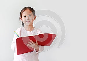 Portrait of little Asian girl writes in a book or notebook with looking beside in classroom against white background with copy