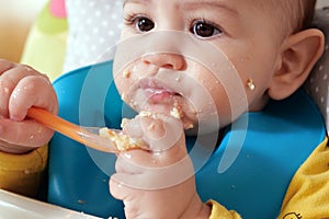Portrait of little baby boy eating food. Baby with a spoon in feeding chair. Cute baby eating first meal