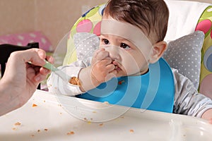 Portrait of little baby boy eating food. Baby with a spoon in feeding chair. Cute baby eating first meal