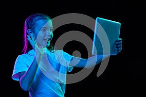 Portrait of little beautiful girl, child in white T-shirt having video call on tablet isolated over black studio