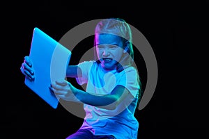 Portrait of little beautiful girl, child in white T-shirt playing on tablet isolated over black studio background in