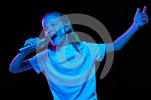 Portrait of little beautiful girl, child in white T-shirt posing, singing in microphone isolated over black studio