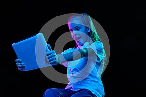 Portrait of little beautiful girl, child in white T-shirt watching cartoon on tablet isolated over black studio