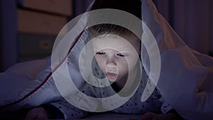 Portrait little blond boy experiences emotion of fear and anger while watching scary movie or cartoon on tablet, lying
