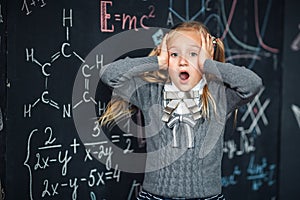 Portrait of little blond girl, grabbing her head. chalkboard with school formulas at the background, concept photo