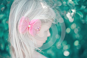 Portrait of little blonde Caucasian girl with pink hair bow. Pretty pensive sad child kid. Girly girl with red hair clip looking