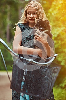 Portrait of a little blonde girl in a casual dress, holds cute spitz dog, in a park.