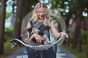 Portrait of a little blonde girl in a casual dress, holds cute spitz dog. Ride on a bicycle in the park.