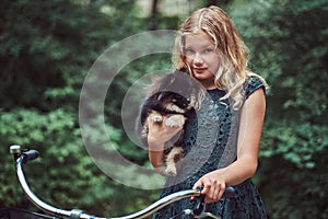 Portrait of a little blonde girl in a casual dress, holds cute spitz dog, in a park.