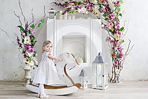 Portrait of a little blonde girl in flowers. She stand near the wooden horse rocker in the white flowers room