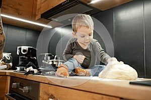 Portrait of little boy bake in kitchen at home. Baby toddler sitting on table with floury legs, touching dough. Cooking.