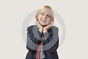 Portrait of little boy with blond hair and blue jacket, keeps hand on cheek, looks directly at camera, isolated over white