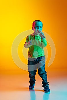 Portrait of little boy in casual style clothes singing at microphone isolated on yellow studio backgroud in neon light