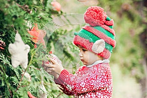 Portrait of a little boy in elf hat and red sweater near the christmas tree and holding decoration