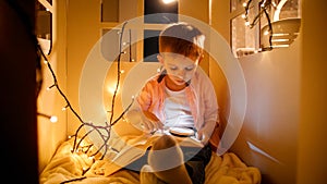 Portrait of little boy with flashlight palying in his toy house at night and reading big book. Concept of child