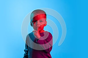 Portrait of little boy, kid, preschool age child isolated on blue studio backgroud in red neon light. Concept of child