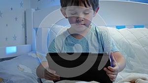 Portrait of little boy lying in bed and playing video games on tablet computer at night. Child with gadget at night