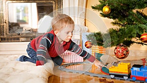 Portrait of little boy sitting on floor at living room and looking at toy train on railways under Christmas tree