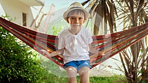 Portrait of little boy sitting in hammock and swaying at garden. Carefree childhood memories, and the relaxation of a