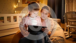 Portrait of little boy sitting on mothers lap and watching cartoons on tablet computer at night. Concept of child