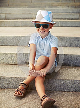 Portrait of little boy in sunglasses and hat sitting and laughing on stairs