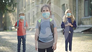 Portrait of little brunette boy in face mask standing in sunlight at school yard with blurred classmates at the