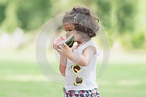 Portrait of little Caucasian cute girl holding sliced watermelon and enjoy eating or biting fresh fruit while picnic at park.
