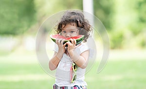 Portrait of little Caucasian cute girl holding sliced watermelon and enjoy eating or biting fresh fruit while picnic at park.